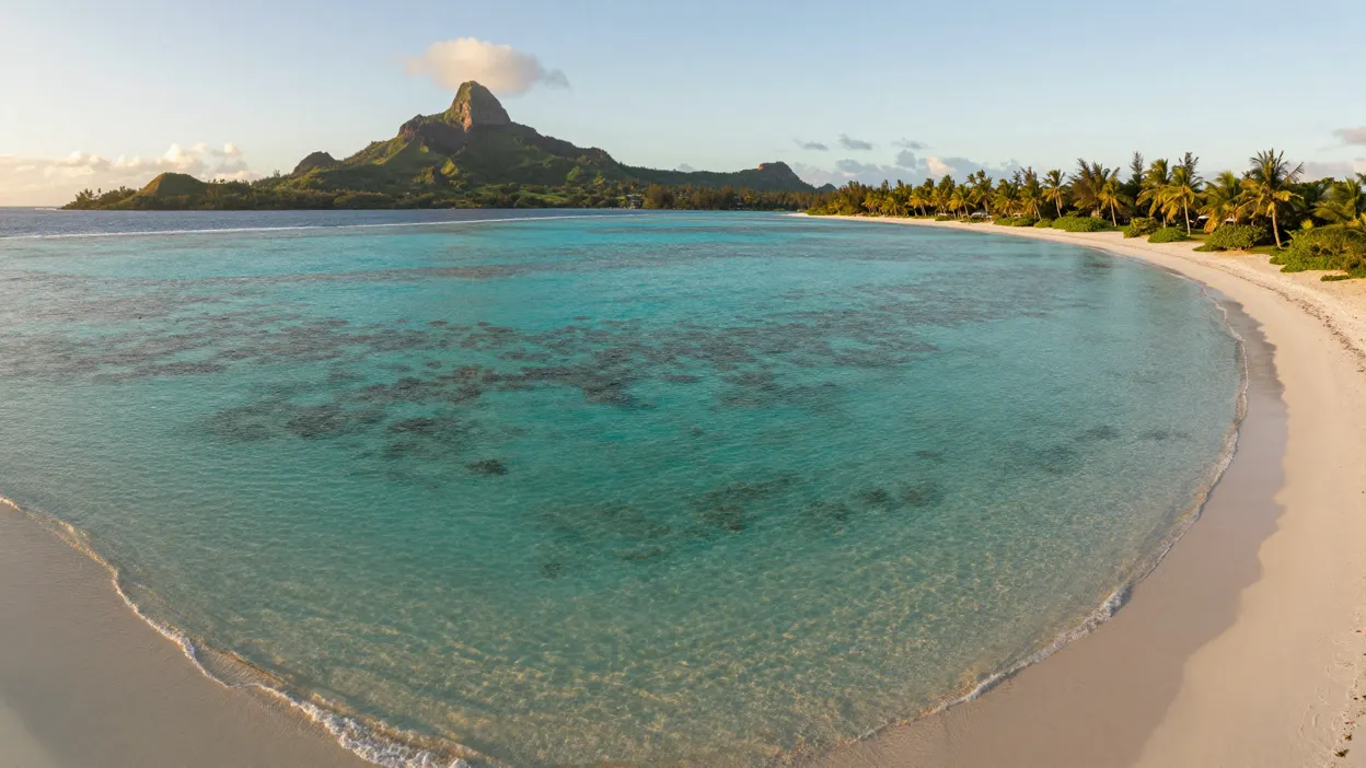 Lagon turquoise de Polynésie au lever du soleil, récif visible sous l'eau claire, pic volcanique vert au fond, plage de sable blanc déserte bordée de cocotiers