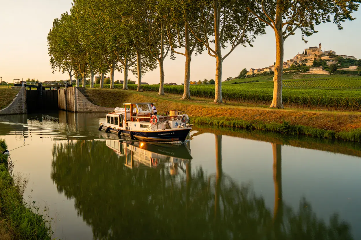 Petit bateau de croisière fluviale glissant sur un canal français au coucher du soleil, écluse de pierre et rangée d'arbres reflétés dans l'eau calme, vignobles au loin
