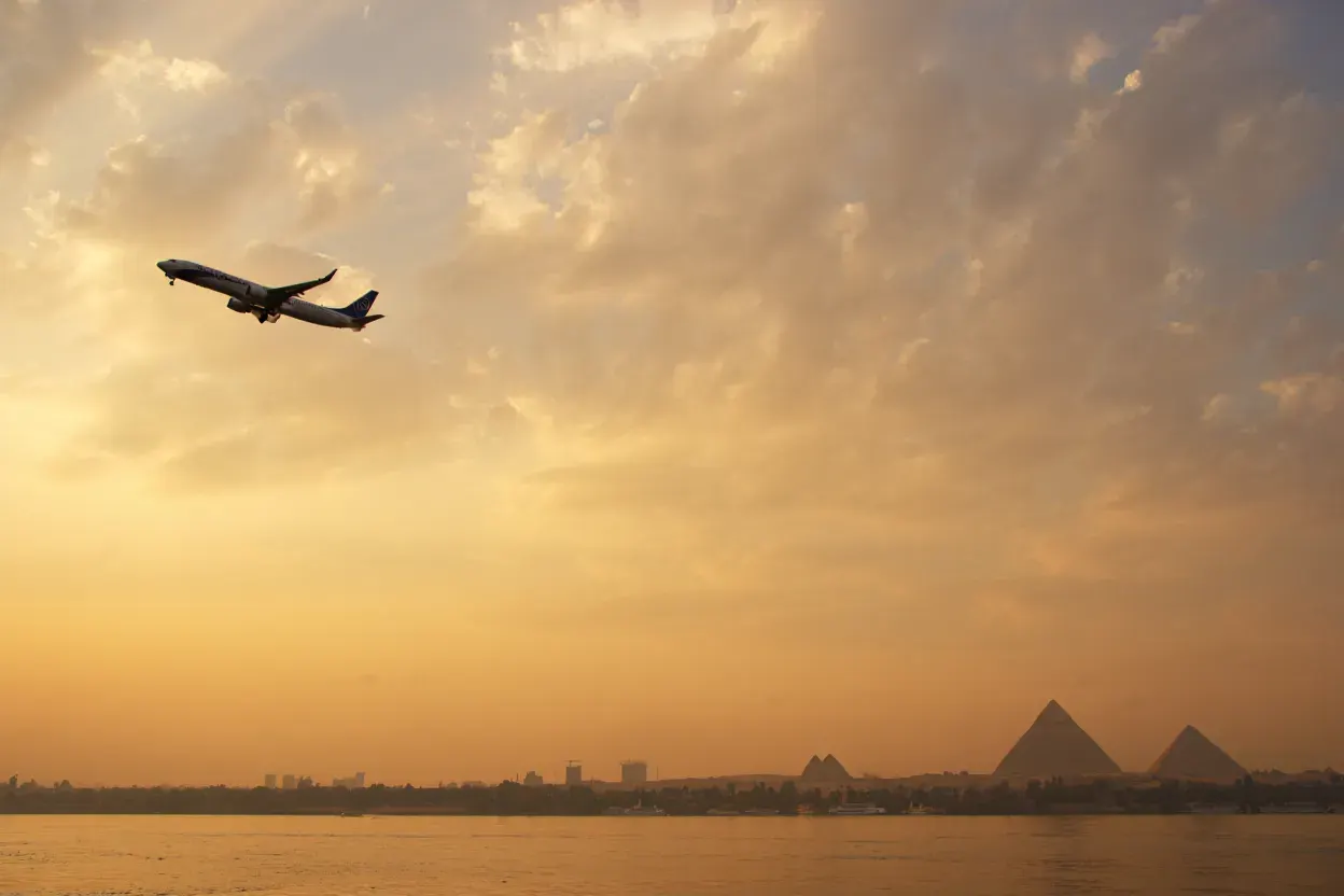 Silhouette d'un avion de ligne traversant un ciel doré au-dessus du Nil et des pyramides de Gizeh dans la brume, au coucher du soleil