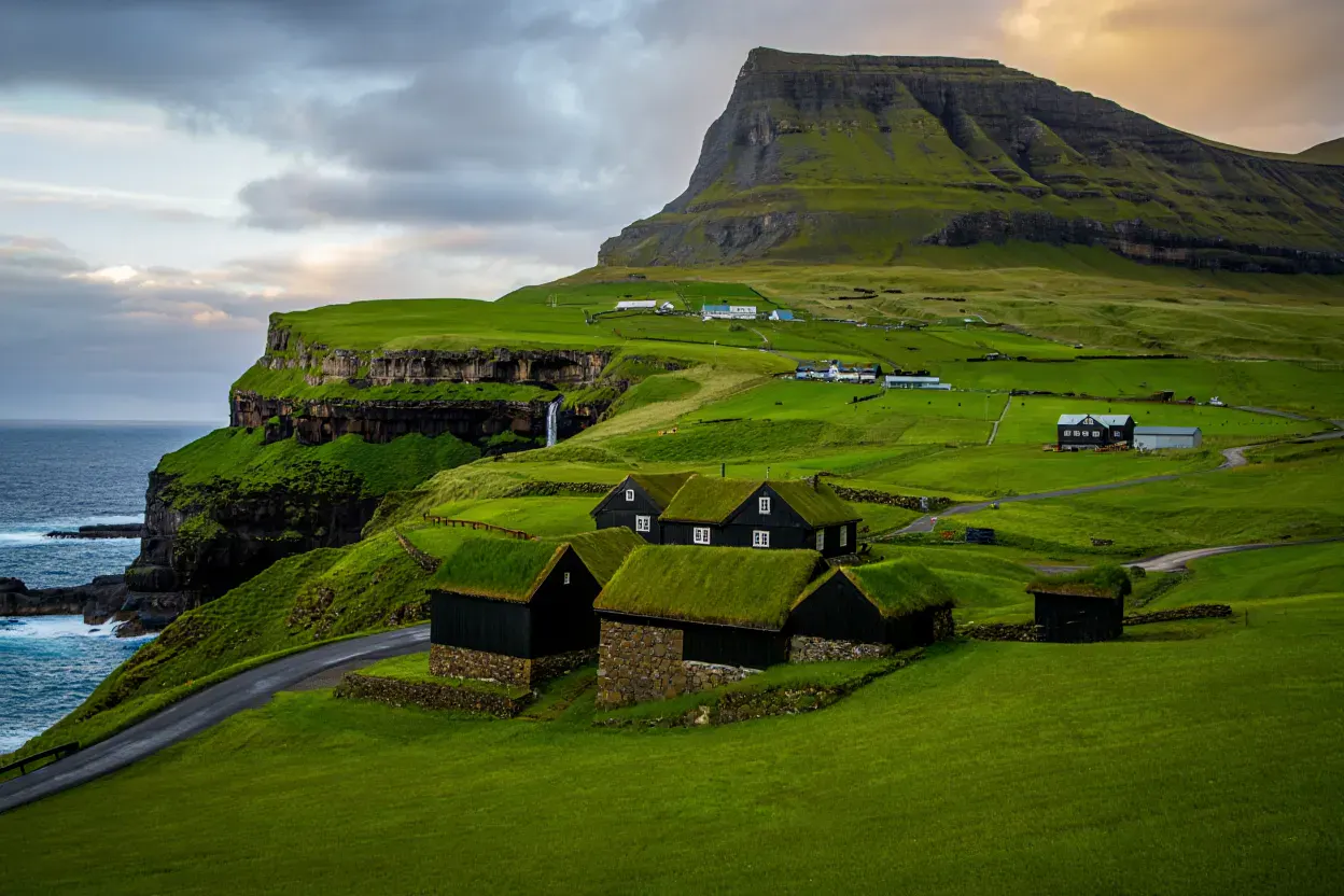 Village féroïen aux maisons à toit d'herbe au pied de falaises vertigineuses, cascade tombant vers l'Atlantique Nord sous un ciel nuageux