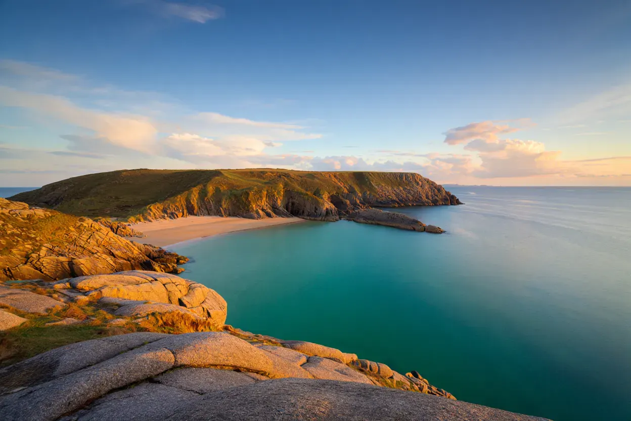 Côte rocheuse de Jersey sous un ciel d'été éclatant, mer turquoise, plage de sable doré bordée de rochers, pas de personnes, lumière chaude de fin de journée