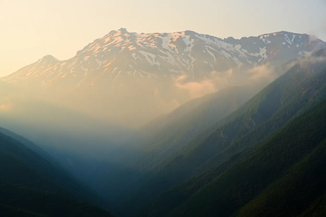 Sommets rocheux et enneigés du mont Olympe en Grèce émergeant de la brume au lever du soleil, pentes boisées vert profond et gorge en contrebas, lumière dorée, aucune présence humaine